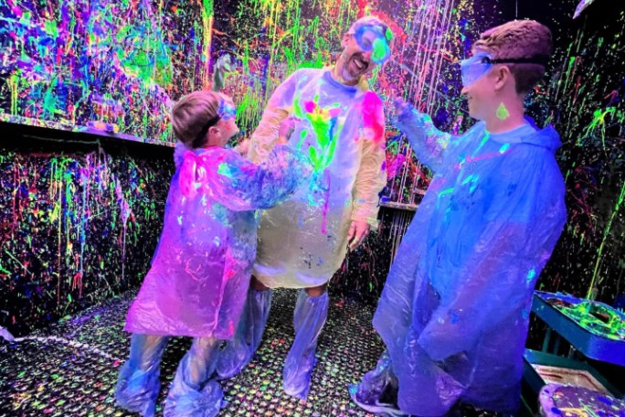 Three people in paint-splattered coveralls enjoying splatter paint room with neon colors.